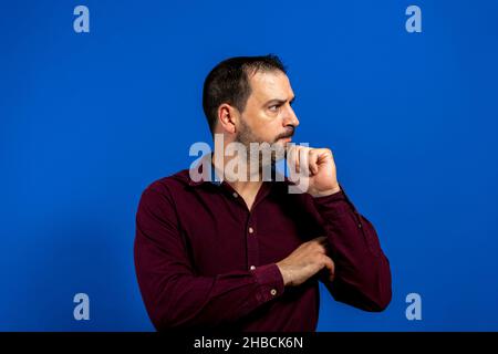 Homme d'affaires hispanique à barbe, portant une chemise violette avec sa main sur le menton, dans un geste réfléchi, isolé sur fond bleu de studio Banque D'Images