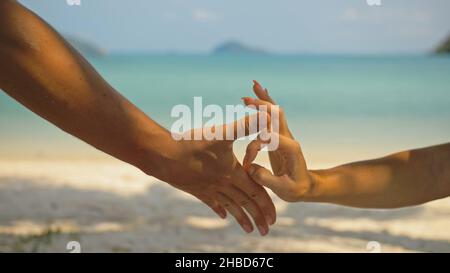 Mains de flirter jeune homme et femme touchant sur la plage dans l'ombre des palmiers contre calme azur océan vue proche. Voyager dans les pays tropicaux Banque D'Images