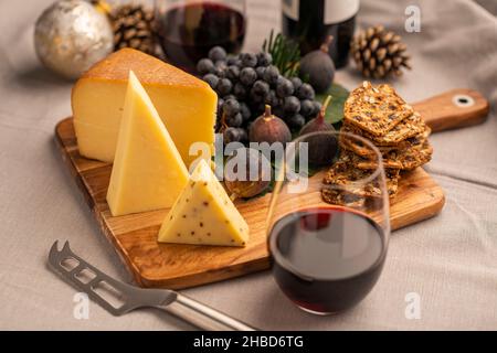 Fête fête fête - planche à fromage avec trois fromages, figues, raisins et craquelins, accompagnés de vin rouge sur une table en tissu. Banque D'Images