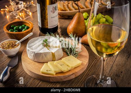 Vin et fromage de fête avec fromage et verre de vin blanc, poires, raisins et olives, lumières scintillantes sur fond de bois. Banque D'Images