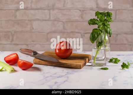 Mains de femme en tablier blanc gants noirs coupant la tomate sur le tableau vert, derrière les légumes frais. Banque D'Images