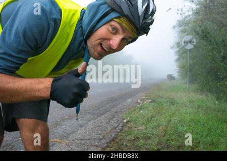 Un cycliste heureux avec une veste réfléchissante vous montre les pouces devant la route dans des conditions brumeuses et un bicycle à proximité.Concepton de vélo sûr sous b Banque D'Images