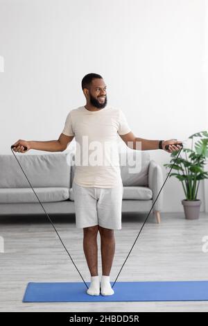 Jeune sportif souriant à barbe noire en vêtements blancs entraînement avec corde à sauter sur tapis dans le salon Banque D'Images