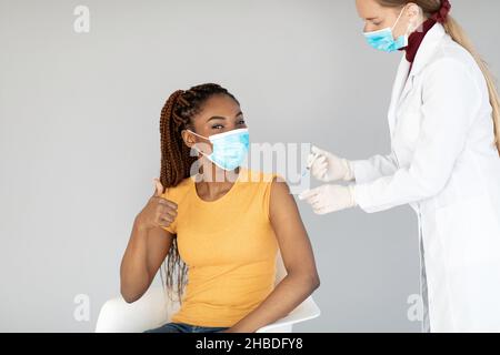 Jeune femme noire en masque facial recevant l'injection de vaccin anti-covid, approuvant la vaccination, en faisant des gestes vers le haut Banque D'Images