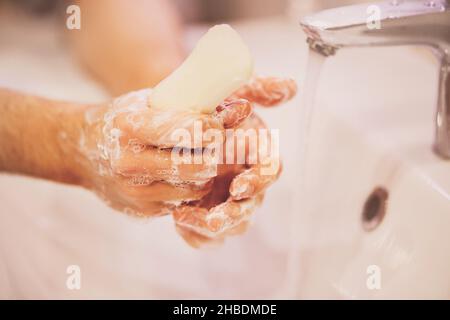 Un homme lave soigneusement ses mains avec du savon sur un lavabo blanc à côté du robinet allumé dans la salle de bains.Propreté et hygiène. Banque D'Images