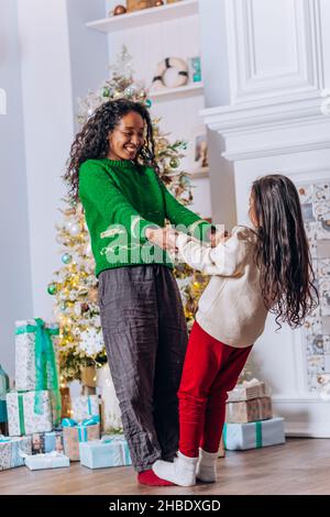 Brunette fille à cheveux longs joue et danse avec une mère afro-américaine souriant joyeusement près de l'arbre de Noël avec des boîtes-cadeaux Banque D'Images