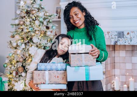 Joyeuse mère afro-américaine et fille brune avec de longs cheveux posent contre les décorations d'arbre de Noël tenant des cadeaux Banque D'Images