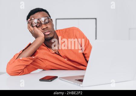 Un jeune homme afro-américain fatigué avec des lunettes dort à portée de main près d'un ordinateur portable à une table blanche dans une salle lumineuse vue rapprochée Banque D'Images