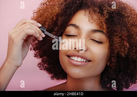 Jeune femme de race mixte avec des cheveux courts bruns et un maquillage parfait tient la pipette et touche le visage avec les yeux fermés gros plan Banque D'Images