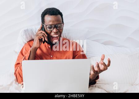 Un homme d'affaires afro-américain en colère, avec des lunettes, criait sur le téléphone de ses subordonnés et regarde l'ordinateur portable qui fonctionne à distance depuis son lit, n'a pas terminé la tâche ou n'a pas respecté la date limite du projet Banque D'Images