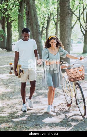 Jeune homme afro-américain avec skateboard et femme brune avec promenade à vélo et sourire dans le parc d'été dans l'après-midi ensoleillé Banque D'Images