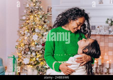 Brunette fille à poil long et la mère maurique afro-américaine posent embrassant et souriant largement contre les décorations d'arbre de Noël Banque D'Images