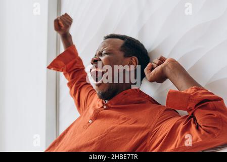 Un jeune homme afro-américain en pyjama orange s'éveille et s'étire dans un lit confortable en début de matinée ensoleillée Banque D'Images