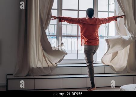 L'homme afro-américain pieds nus en chemise orange lâche ouvre des rideaux regardant par la fenêtre dans la chambre lumineuse et spacieuse vue arrière Banque D'Images