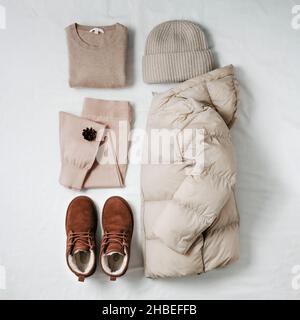 Veste en duvet léger à motif de bourreur chaud pour femmes, chandail en cachemire beige, jogging tricoté rose, chapeau bini tricoté, bottes en daim Chukka en fourrure marron sur drap de lit blanc.Vêtements d'hiver confortables, coloris beige monochrome Banque D'Images