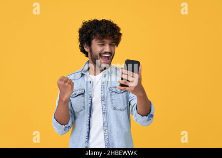 Jeune Indien gagnant enthousiaste utilisant un smartphone isolé sur fond jaune. Banque D'Images