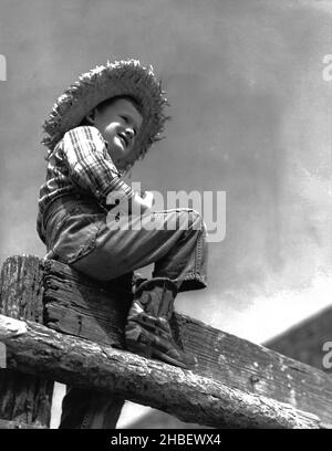 Jeune garçon de ferme dans des bottes de cowboy et chapeau de paille assis sur la vieille clôture en bois regarder dehors.Vers 1962 Banque D'Images