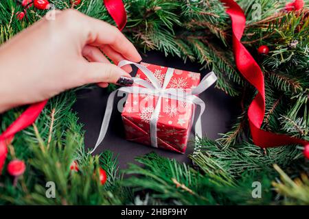 Une femme ouvre une boîte cadeau de Noël ou du nouvel an emballée dans un papier et un ruban de fête à l'intérieur d'une couronne de sapin.Cadeau pour les vacances d'hiver Banque D'Images