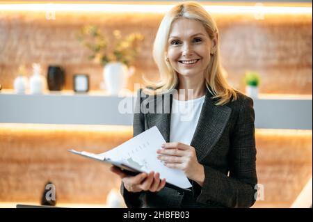 Femme d'affaires ou directrice d'âge caucasien, élégante et réussie, se tient près du bureau, portant un costume habillé élégant, tient des documents financiers, regarde l'appareil photo, sourit amical Banque D'Images