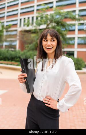 Jeune femme d'affaires caucasienne souriante avec presse-papiers se sentant heureuse après avoir réussi une affaire avec un client ou un partenaire étranger tout en se tenant debout dans la rue de la ville avec une tenue formelle.Concept Business People Banque D'Images