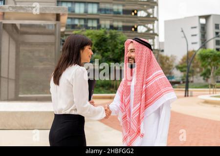 Homme arabe souriant dans des vêtements traditionnels se serrant la main avec une femme d'affaires européenne tout en se tenant debout dans la rue à l'extérieur de la ville.Concept de réunion d'affaires multiraciale Banque D'Images