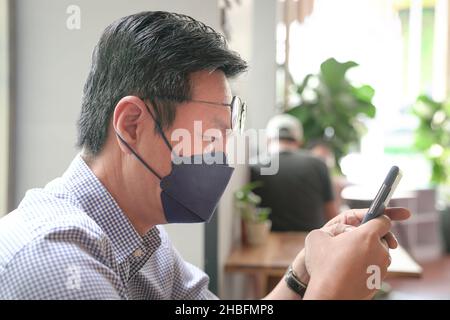 Homme asiatique avec masque sur l'utilisation de son téléphone, messagerie texte dans un restaurant. Banque D'Images