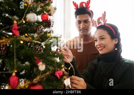 Joyeux jeune couple dévorant l'arbre de Noël à la maison avec des boules colorées Banque D'Images