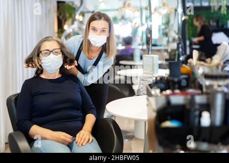 Coiffeur professionnel en masque discutant de coupe de cheveux avec une cliente âgée Banque D'Images