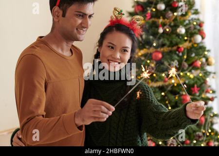 Joyeux jeune couple divers brûlant des lumières du bengale à la fête de Noël à la maison Banque D'Images