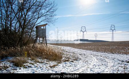Tour de chasse en bois près du champ agricole en hiver.Siège Huntsman haut avec lignes haute tension et pylônes de puissance. Banque D'Images