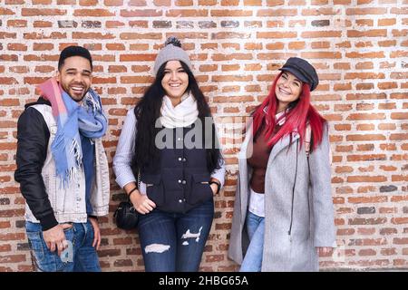 Jeunes amis multiraciaux positifs, hommes et femmes, vêtus de tenues décontractées à la mode, debout près du mur de briques et souriant à l'appareil photo Banque D'Images