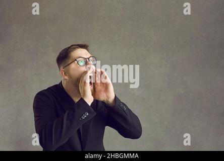 L'homme émotionnel se tient sur un fond gris et criait bruyamment, en tenant ses mains à sa bouche. Banque D'Images