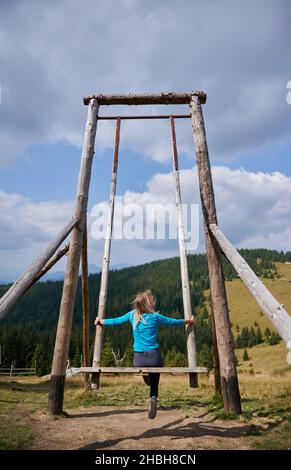 Vue arrière de la jeune femme balançant sur une énorme balançoire en bois mettant les mains sur les supports latéraux au milieu de la pelouse extérieure en été, en admirant le paysage environnant. Banque D'Images