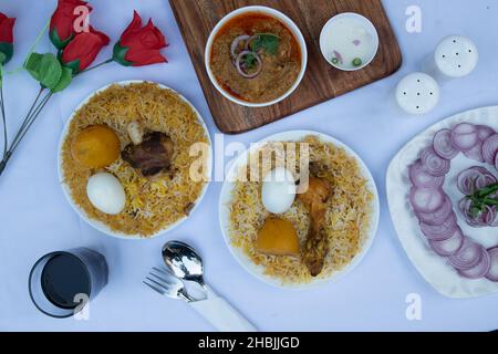 Dose de poulet biryani dans deux assiettes avec des oignons en tranches et jus de fruits avec des roses rouges sur la table blanche propre Banque D'Images