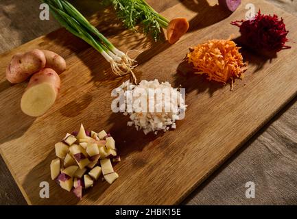 Betteraves déchiquetées, carottes, oignons et cubes de pommes de terre coupées sur une planche de bois avec des moitiés de légumes avec des feuilles vertes et des racines sur le fond.Fermer- Banque D'Images