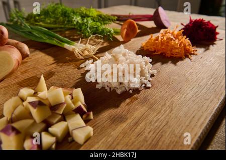 Gros plan de betteraves râpées, carottes, oignons et cubes de pommes de terre coupées sur une planche de bois avec des moitiés de légumes avec des feuilles vertes et des racines sur le Banque D'Images