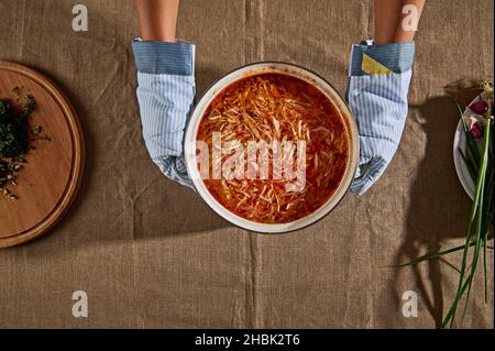 Vue en hauteur des mains du chef dans des gants de cuisine, en tenant une casserole de soupe de borscht fraîchement préparée et en la plaçant sur une table avec une nappe en lin à côté de cuttin Banque D'Images