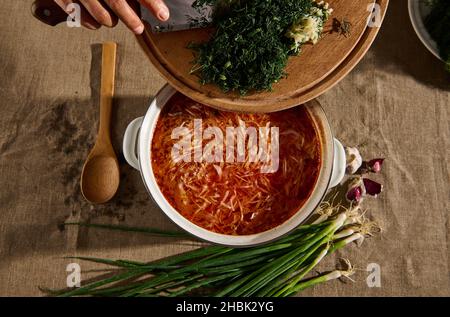 Vue de dessus: Les mains du chef verser l'aneth frais haché et l'ail dans une casserole de soupe ukrainienne traditionnelle maison fraîchement préparée - borscht.Ukraine Banque D'Images