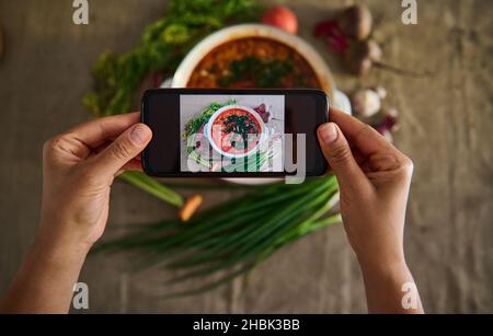 Vue de dessus des mains tenant un smartphone et photographier un pot avec une soupe traditionnelle ukrainienne fraîchement cuite - Borscht avec de la verdure couché sur une ta de lin Banque D'Images