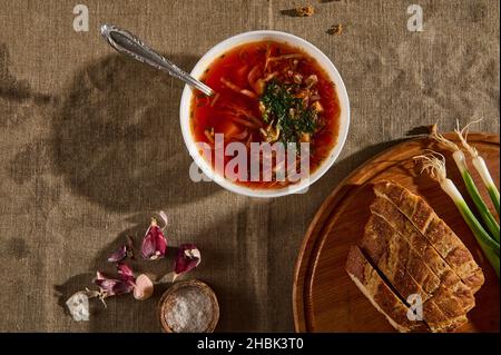 Bol en céramique blanche avec borscht, oignons verts et pain tranché sur planche à découper, ail pelé et contenant de sel sur nappe en lin.Plat Banque D'Images