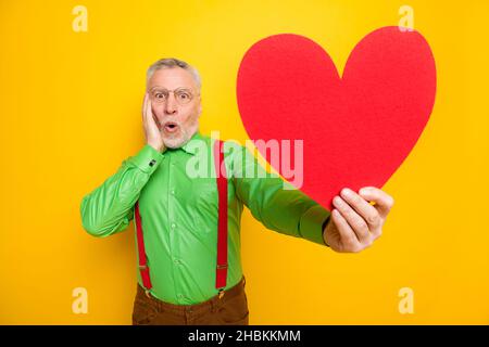 Photo portrait homme aîné stupéfié démontrant rouge coeur en forme de carte postale sur date isolé couleur jaune vif fond Banque D'Images