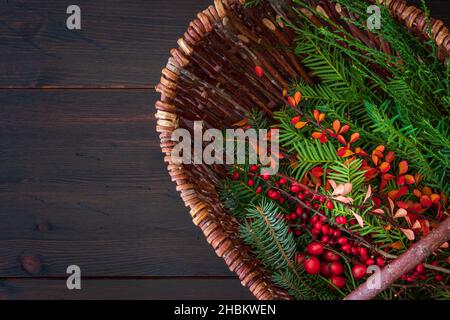 Sélection de branches d'hiver dans un panier en osier sur fond de bois rustique brun foncé.Branches de sapin vert et d'if avec hanches roses.Arrière-plan d'hiver. Banque D'Images