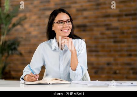 Pensive positive SMART jeune adulte brune femme d'affaires caucasienne, gestionnaire ou courtier portant des lunettes, assis à table au bureau, regardant à côté, prenant une pause de travail, rêvant de vacances Banque D'Images