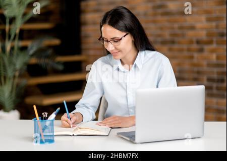 Smart attrayant succès jeune caucasien brune femme d'affaires ou courtier assis à son bureau étudiant ou travaillant sur un ordinateur portable à l'écoute de conférences en ligne, webinar, notes d'information Banque D'Images