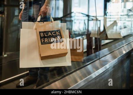 Photo courte d'un homme méconnaissable tenant des sacs à provisions avec une affiche de solde du Vendredi fou en se tenant à l'escalier roulant du centre commercial, dans l'espace réservé aux copies Banque D'Images