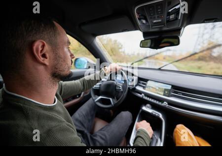 Vue rapprochée du conducteur derrière la roue de la voiture.Jeune homme barbu assis confortablement sur le siège du conducteur, en plaçant une main sur le volant, l'autre sur la boîte de vitesses et en regardant la route devant vous. Banque D'Images