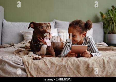 Vue de face portrait d'une jolie fille afro-américaine couchée sur un lit avec un grand chien d'animal de compagnie et souriant, espace de copie Banque D'Images