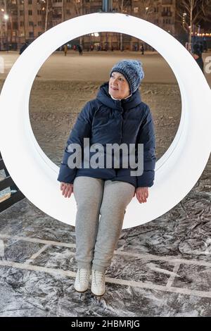 Portrait d'une femme âgée en patins à glace qui se repose sur un banc à anneau à la patinoire de la ville en soirée d'hiver. Banque D'Images