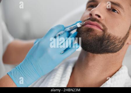 L'homme attrayant avec la barbe subit une procédure d'augmentation de la lèvre avec le cosmétologue en clinique Banque D'Images