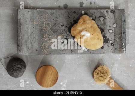 Éponge de mer biologique avec mousse de savon sur plateau en béton gris, crème pour le visage dans un pot avec couvercle en bois, éponge konjac de charbon noir et brosse pour le visage.Accès à la salle de bains Banque D'Images